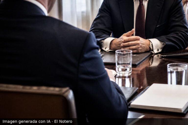 Dos delegados anónimos sentados frente a una mesa de reuniones, con carpetas y vasos de agua, en una sala iluminada por la luz natural de una ventana.