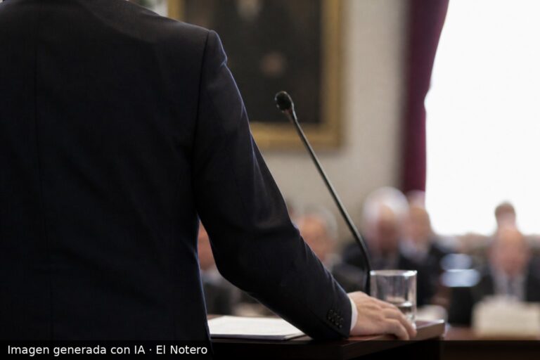 Un economista de espaldas habla en un atril dentro de una sala institucional, con asistentes desenfocados al fondo y luz natural entrando por una ventana.