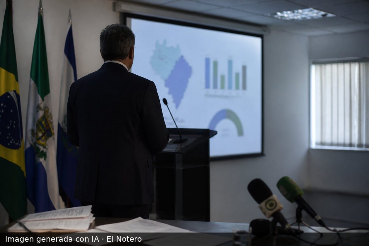 Una sala de prensa institucional con un funcionario de espaldas junto a una pantalla con gráficos de resultados electorales desenfocados y micrófonos en primer plano.