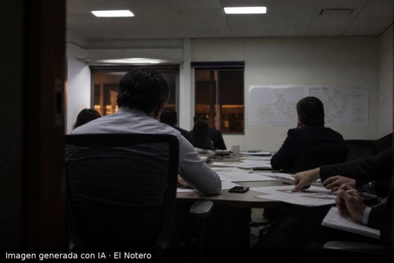 Sala de reuniones con personas sentadas alrededor de una mesa, documentos electorales sobre la mesa y una atmósfera tensa, vistos desde el fondo sin rostros visibles.