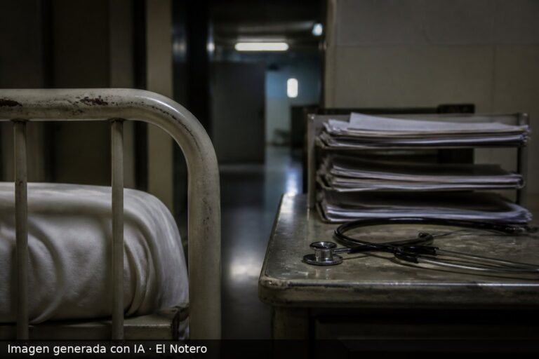 Una cama metálica de cárcel junto a formularios médicos apilados y un estetoscopio sobre una mesa gastada, en un ambiente institucional de luz fría.