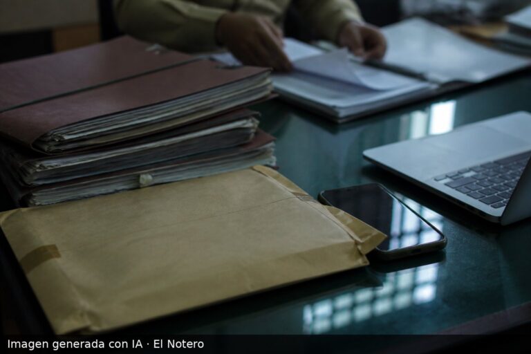 Primer plano de expedientes, un sobre cerrado, un teléfono y una computadora portátil sobre un escritorio oficial, con manos desenfocadas ordenando papeles al fondo.