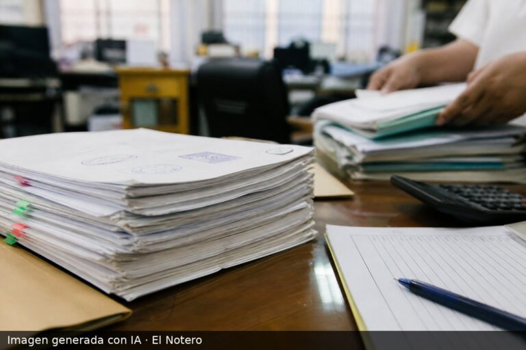 Una pila de expedientes y contratos administrativos sobre un escritorio, con una oficina municipal desenfocada al fondo y manos ordenando papeles.