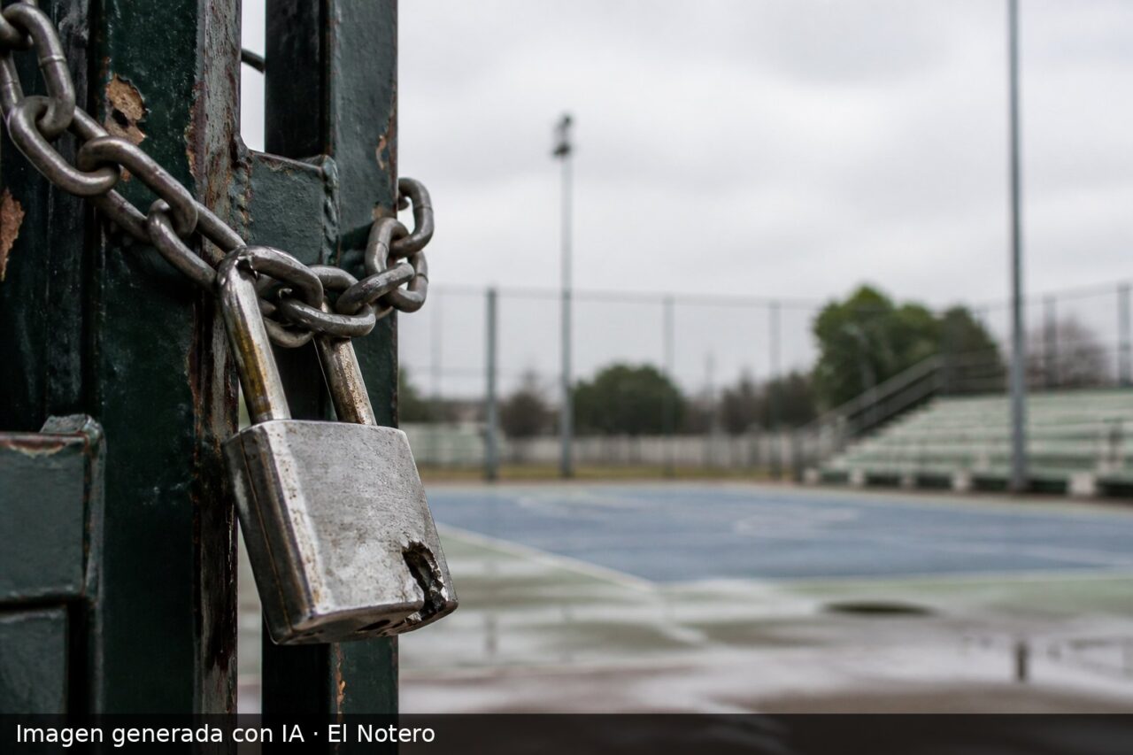 Primer plano de un candado dañado en el acceso a un polideportivo municipal, con la cancha y las tribunas vacías desenfocadas al fondo.
